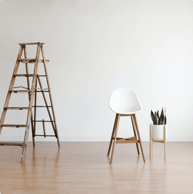 Holzleiter, weißer Stuhl und Pflanze in minimalistisch eingerichtetem Raum mit hellem Holzboden und weißer Wand.